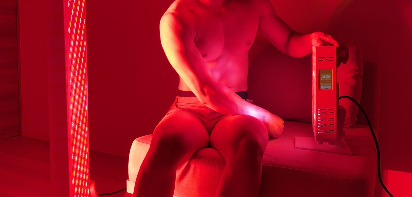 赤い光に照らされた部屋で、運営者がソファに座り、卓上型レッドライトパネルのタイマーを操作しているようす。左には大型のスタンド型パネルが点灯している。